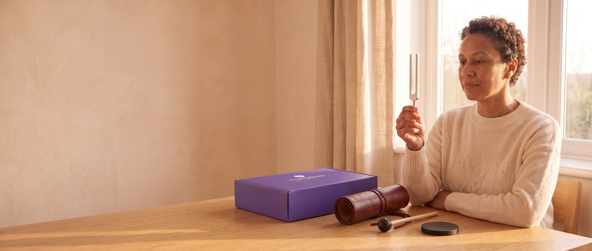 A woman at a sunny table holds up a tuning fork, the Tranquitone gift box, a rolled-up case, a mallet and a puck on the table.