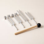 Four tuning forks, two weighted and two plain, and a wooden mallet on warm paper.