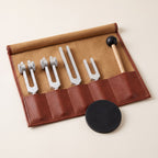 An open roll-up case with four tuning forks and a wooden mallet, and a black striking puck in front.