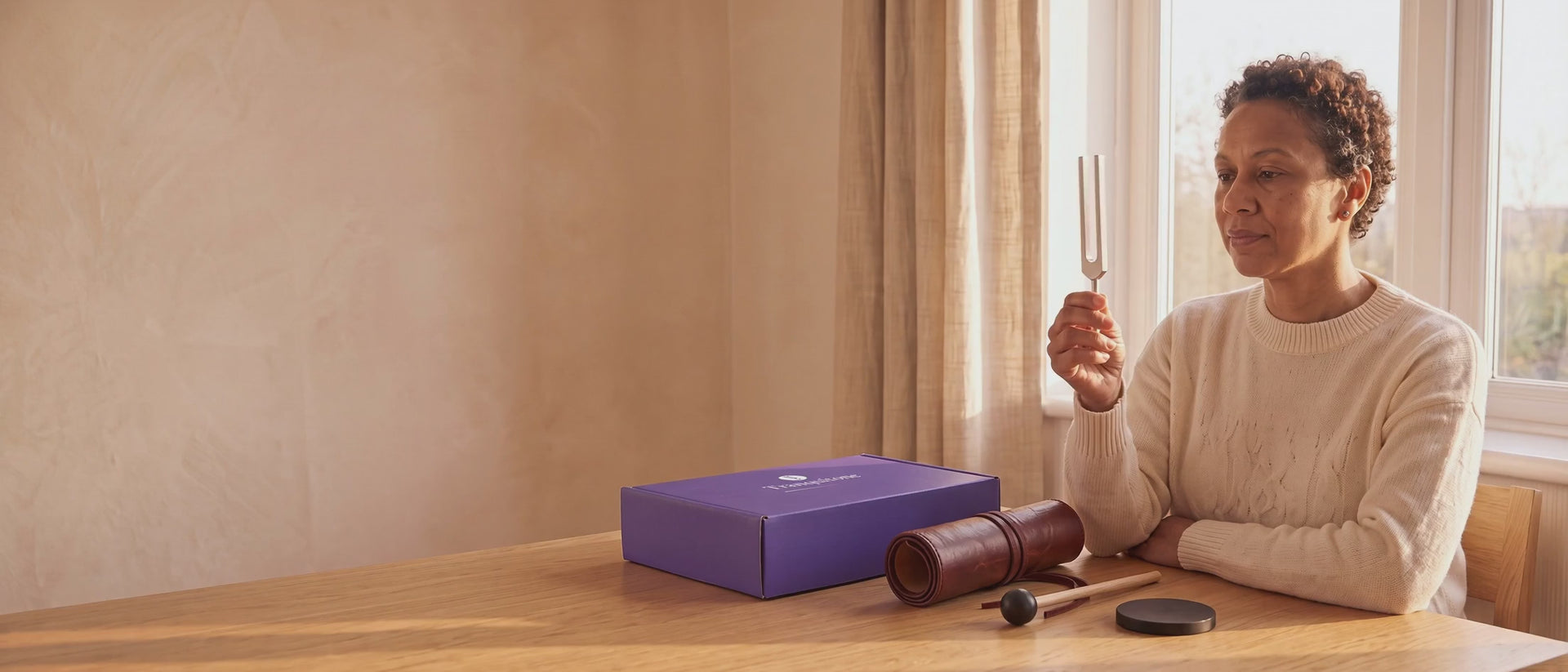 A woman at a sunny table holds up a tuning fork, the Tranquitone gift box, a rolled-up case, a mallet and a puck on the table.
