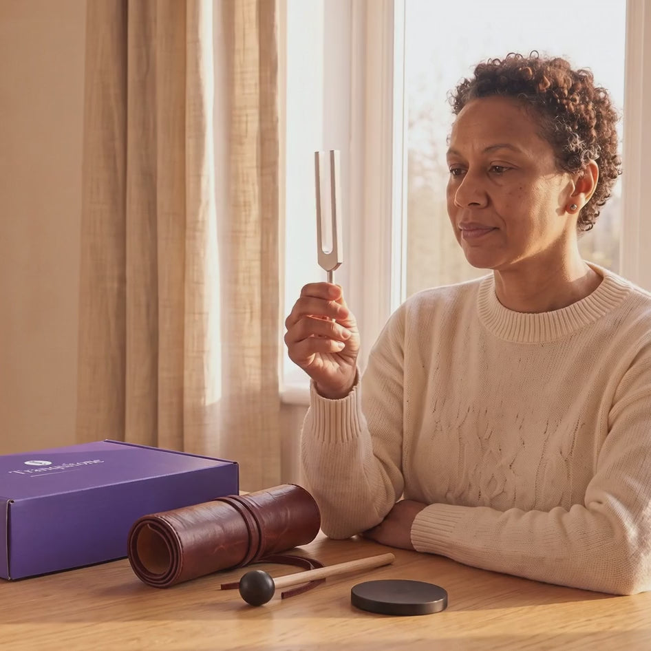 A woman at a sunny table holds up a tuning fork, the Tranquitone gift box, a rolled-up case, a mallet and a puck on the table.