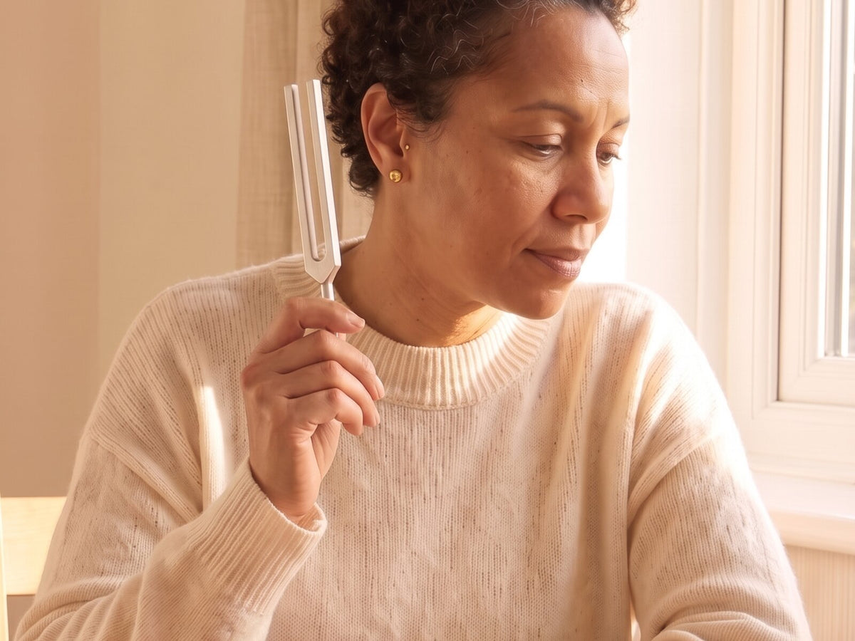 A woman holds a tuning fork upright beside her ear, with a gap between the fork and her ear.